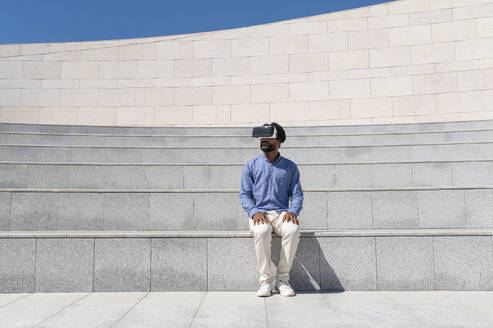 Junger Mann mit Virtual-Reality-Simulator auf einer Treppe sitzend - VRAF00211