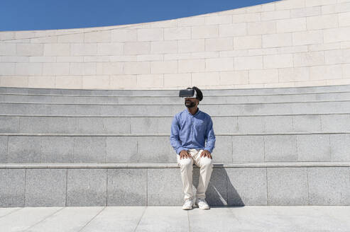 Young man wearing virtual reality simulator sitting on staircase - VRAF00211