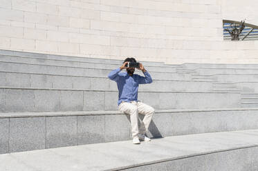 Junger Mann sitzt auf einer Treppe und trägt einen Virtual-Reality-Simulator - VRAF00212
