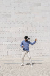 Junger Mann mit Virtual-Reality-Simulator gestikuliert vor einer Wand - VRAF00215