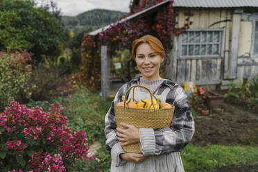 Smiling redhead farmer carrying basket of pumpkins at farm - VBUF00430