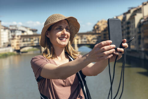 Smiling tourist wearing hat taking selfie with Ponte Vecchio over Arno River in background - JSRF02650