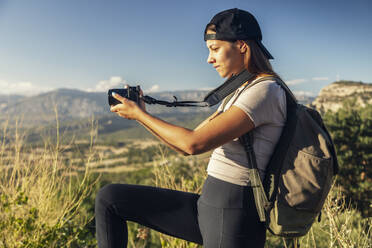 Junge Frau mit Rucksack beim Fotografieren an einem sonnigen Tag - JSRF02654