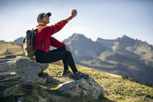 Young woman sitting on rock taking selfie through smart phone - JSRF02668