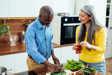 Cinematic image of a multiethnic senior couple preparing food in the kitchen. Indoors Lifestyle moments at home. Concept about seniority and relationships - DMDF07586