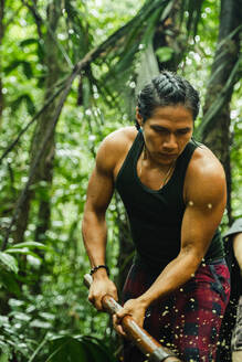 Fokussierter ethnischer Mann mit geflochtenem Haar, der mit einem Stock arbeitet, während er im üppigen Grün des Amazonas-Regenwaldes steht - ADSF48027