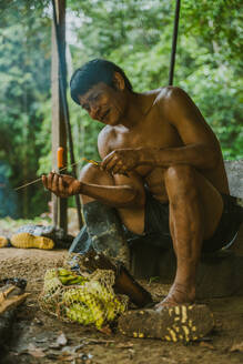 Ganzkörperdarstellung eines ethnischen Huaorani-Mannes, der unter einem Schuppen in einem Dorf im Amazonas-Regenwald sitzt und Curare-Pfeile für die Jagd herstellt - ADSF48030