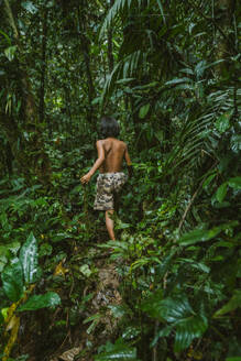 Rückenansicht eines anonymen Teenagers in Shorts, der langsam auf einem Hang im üppigen grünen Gebüsch des Amazonas-Regenwaldes läuft - ADSF48031