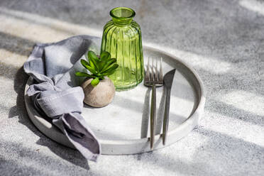 High angle of minimalist table setting with small potted plant on plate with cutlery and empty bottle in sunlight - ADSF48040