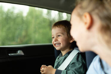 Side view of children having fun and laughing while sitting in vehicle with fastened seat belt in daylight against blurred trees - ADSF48042