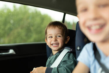 Children having fun and laughing while sitting in vehicle with fastened seat belt in daylight against blurred trees - ADSF48043