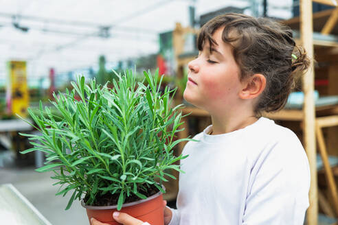 Side view of preteen girl with closed eyes while standing and carrying potted green leaves rosemary herbal plant and smelling fragrance - ADSF48049