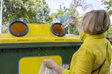 Back view of anonymous mature blond woman throwing plastic bottles from reusable bag into yellow recyclable trash against blurred trees - ADSF48065