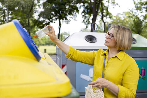 Side view of mature blond woman with eyeglasses throwing plastic bottles from reusable bag into yellow recyclable trash against blurred trees and glass container - ADSF48068