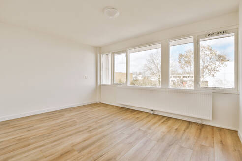 Interior of empty living room with radiator on white wall by large bright windows and parquet floor in contemporary apartment - ADSF48084
