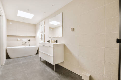 Interior of spacious illuminated bathroom with bathtub in corner and mirror with sink and cabinet by tiled wall in modern apartment - ADSF48085