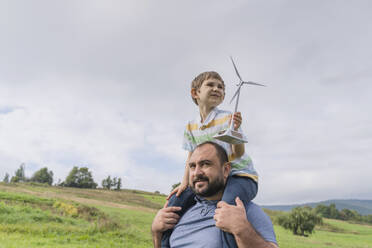 Father carrying son on shoulders with wind turbine model in front of sky - OSF02278