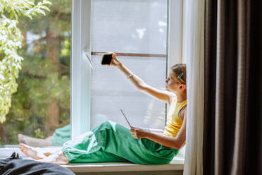 Girl taking selfie over smart phone sitting on window sill - MDOF01592