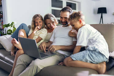 Man sharing laptop with family sitting on sofa at home - JSRF02703