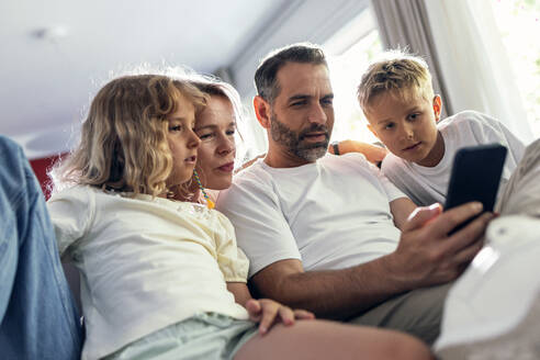 Vater teilt Mobiltelefon mit Familie im Wohnzimmer sitzend - JSRF02704