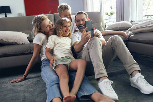 Smiling man taking selfie with family on smart phone in living room - JSRF02707