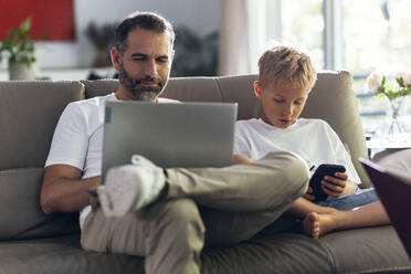 Father working over laptop by son using smart phone on sofa - JSRF02712