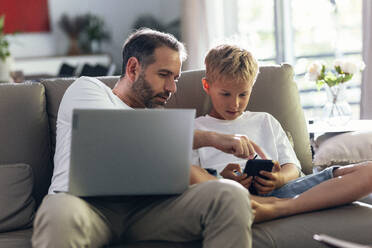 Vater und Sohn benutzen ein Mobiltelefon auf dem Sofa zu Hause - JSRF02713