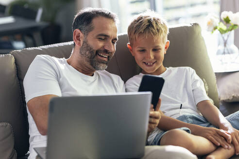 Lächelnder Vater teilt Smartphone mit Sohn im Wohnzimmer - JSRF02714