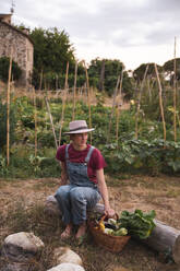 Frau in Latzhose sitzt mit einem Korb voller Gemüse in einem Obstgarten - PCLF00809