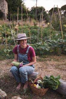 Lächelnde Frau auf einem Baumstamm sitzend mit einem Korb voller Gemüse im Obstgarten - PCLF00811