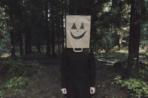 Woman covering face with paper bag standing in forest - VBUF00452