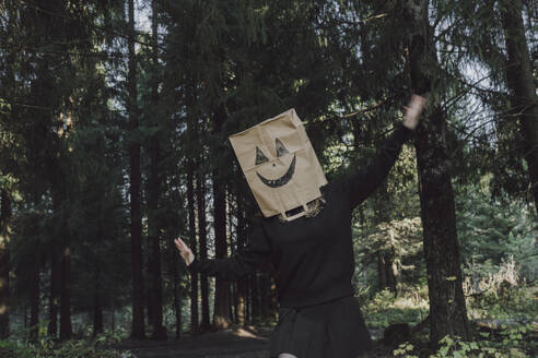 Unbeschwerte Frau trägt Papiertüte über dem Gesicht und tanzt im Wald - VBUF00454