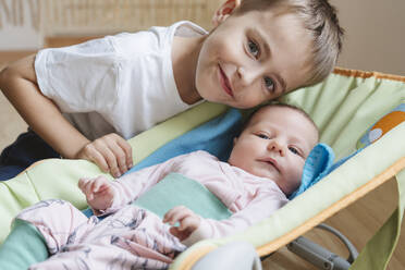 Lächelnder Junge neben seiner neugeborenen Schwester, die zu Hause in der Babywippe liegt - IHF01757
