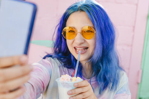 Happy woman with dyed blue hair holding drink taking selfie at cafe - YTF01237