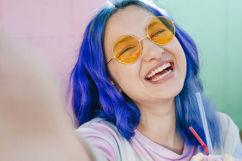 Happy hipster woman with dyed blue hair holding drink taking selfie at cafe - YTF01239
