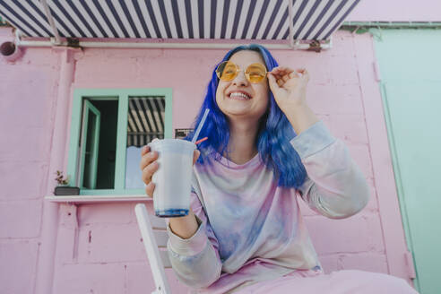 Glückliche Frau mit gefärbten blauen Haaren hält Milchshake in einem Café - YTF01240