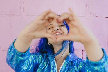 Lächelnde Frau mit blau gefärbtem Haar schaut durch eine Herzform - YTF01248