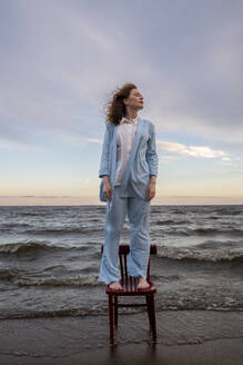 Young woman standing on chair in front of sea - VPIF08907