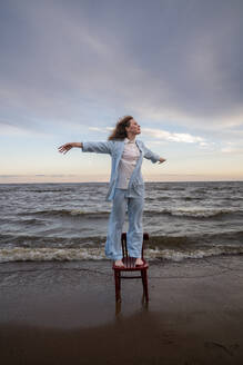 Young woman standing with arms outstretched on chair at beach - VPIF08908