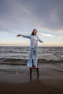 Woman standing with arms outstretched on chair at beach - VPIF08909