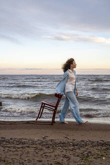 Junge Frau beim Spaziergang mit Stuhl am Strand - VPIF08915