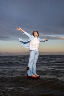 Young woman standing on chair in sea with arms outstretched under cloudy sky - VPIF08921