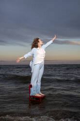 Young woman standing on chair in sea with arms raised under cloudy sky - VPIF08922