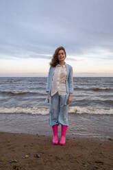 Junge Frau in rosa Gummistiefeln steht am Strand - VPIF08924