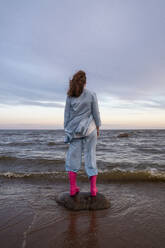 Junge Frau in rosa Gummistiefeln steht auf einem Felsen am Strand - VPIF08925