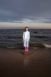 Woman wearing pink rubber boots standing on rock at sunset - VPIF08928