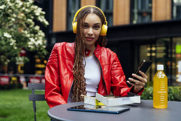 Frau mit Mobiltelefon hört Musik über Kopfhörer am Tisch in einem Straßencafé - OIPF03520