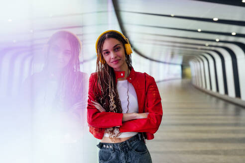 Smiling woman with arms crossed listening music on headphones in underground tunnel - OIPF03528