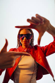 Junge Frau mit Sonnenbrille gestikuliert Fingerrahmen vor weißer Wand - OIPF03541