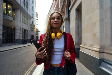 Smiling woman walking with smart phone and backpack on street in city - OIPF03559